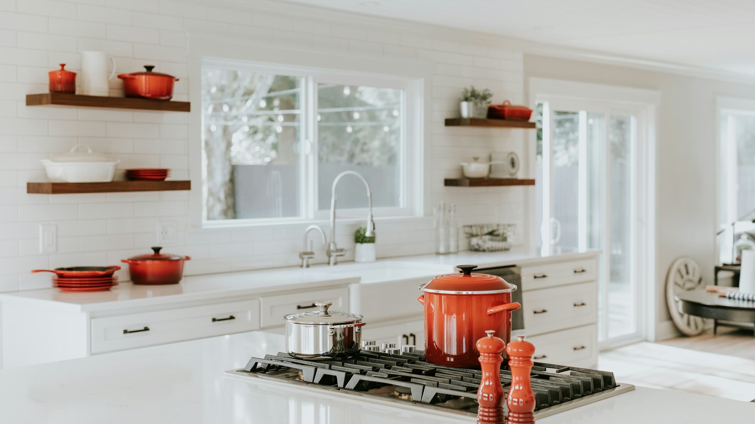 A bright white kitchen with a red enameled stockpot on the gas stove and red cookware on open wooden shelves